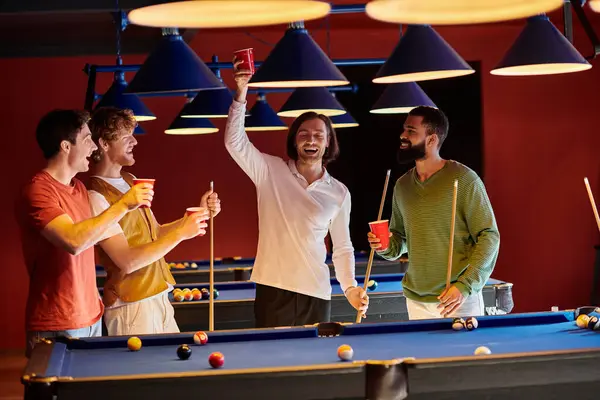 Gli amici festeggiano una vittoria durante una partita di biliardo casuale, ridendo e alzando le coppe rosse in aria. — Foto stock