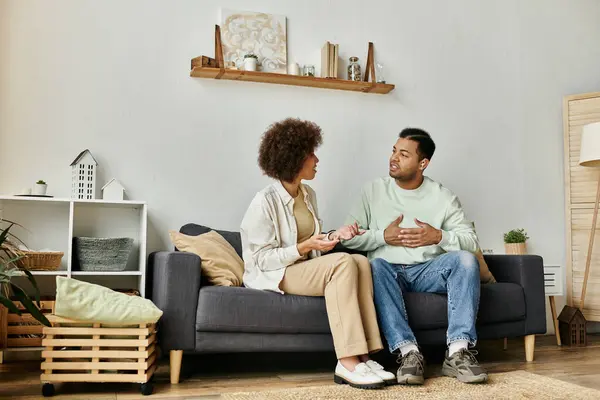 Una atractiva pareja afroamericana se sienta en un sofá en su sala de estar, usando lenguaje de señas para comunicarse. - foto de stock