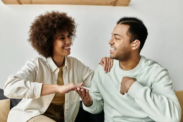 Афроамериканская пара общается через язык жестов, улыбаясь и смеясь, сидя на диване в своем доме. — стоковое фото