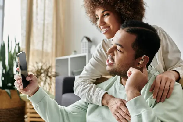 Joyous African American woman sitting near her husband with hearing aid and looking at phone. — Stock Photo