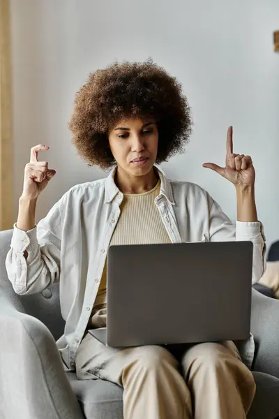 A woman with curly hair sits on a couch and uses sign language while looking at a laptop. — Stock Photo