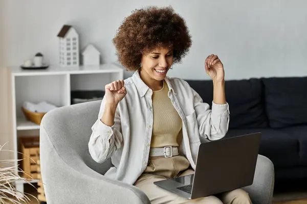 Une jeune femme afro-américaine attrayante s'assoit sur une chaise, souriant largement et avec enthousiasme en utilisant le langage des signes, à côté de l'ordinateur portable. — Photo de stock