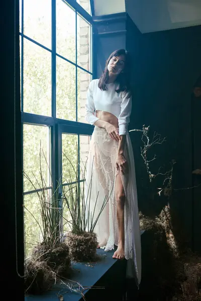 A woman in a white top and sheer skirt stands near a window with green plants, gazing out. — Stock Photo