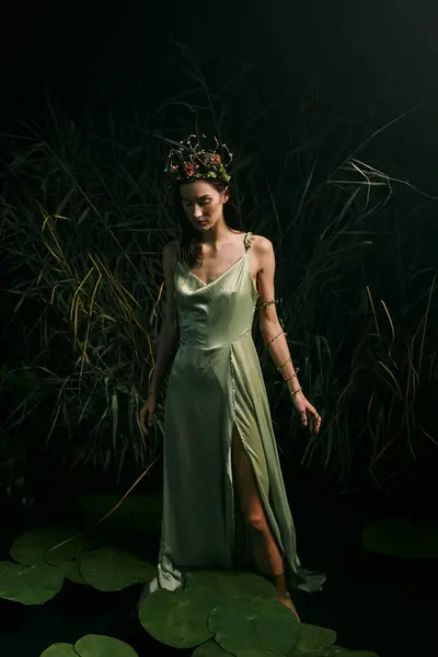 A woman stands gracefully on lily pads in a swamp, wearing a flowing green dress and a floral crown. — Stock Photo