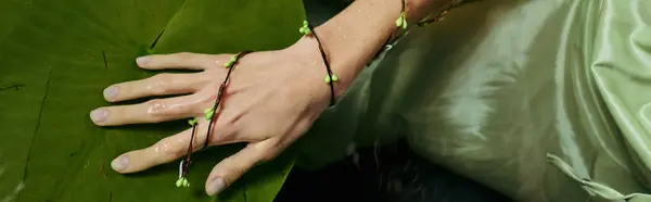 Una mano femminile con gioielli floreali verdi poggia su una grande foglia verde vicino a una palude. — Foto stock