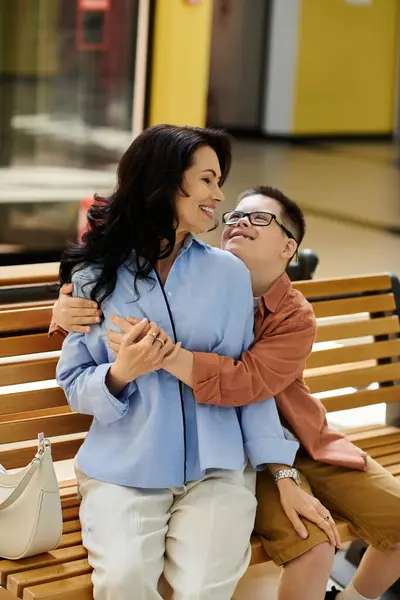 A mother and her son with Down syndrome share a warm embrace while sitting on a bench in a shopping mall. — Stock Photo