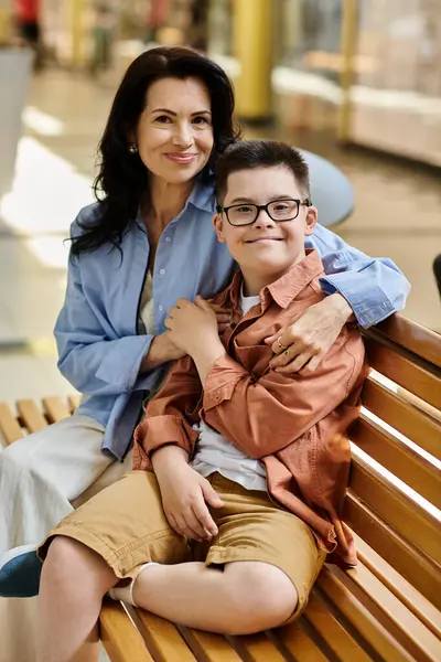 A mother and her son with Down syndrome sit on a bench in a shopping mall, smiling for the camera. — Stock Photo