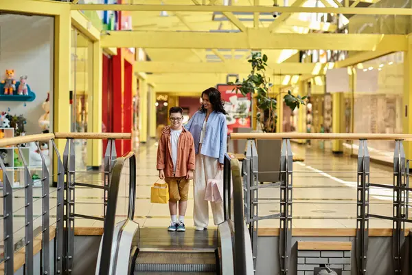 A mother and her son with Down syndrome are smiling while riding an escalator at a shopping mall. — Stock Photo