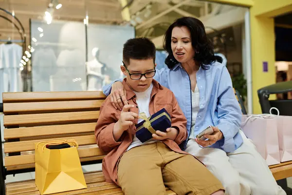 A mother and her son with Down syndrome sit on a bench in a shopping mall, sharing a gift and a warm moment together. — Stock Photo
