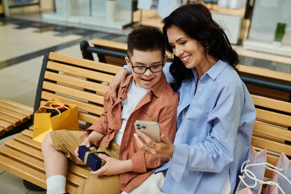 A mother and her son with Down syndrome smiling together, looking at a phone on a mall bench. — Stock Photo