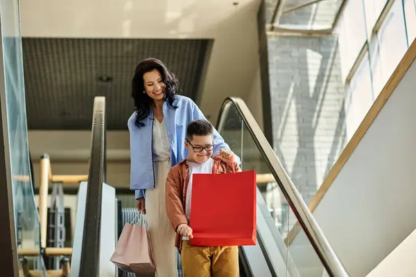 Uma mãe e seu filho com síndrome de Down desfrutam de um dia de compras no shopping, andando na escada rolante e carregando sacos de compras. — Fotografia de Stock