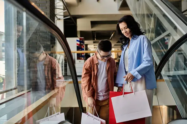 Una madre e suo figlio con la sindrome di Down camminano lungo una scala mobile in un centro commerciale, entrambi con borse della spesa. — Foto stock