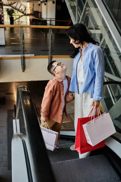 Una madre e suo figlio con la sindrome di Down godono di una giornata di shopping al centro commerciale, camminando lungo la scala mobile insieme. — Foto stock
