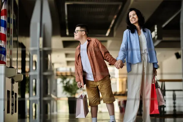 A mother and her son with Down syndrome walk hand-in-hand through a shopping mall, carrying shopping bags. — Stock Photo