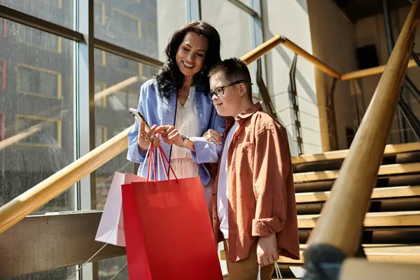 Una madre y su hijo con síndrome de Down están de compras juntos en un centro comercial, llevando bolsas y mirando un teléfono inteligente. - foto de stock