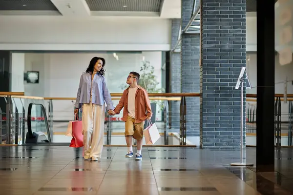 Una madre e suo figlio con la sindrome di Down camminano mano nella mano attraverso un centro commerciale, portando borse della spesa.. — Foto stock