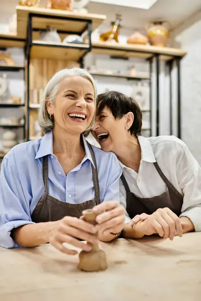 Two mature women laugh together in a pottery studio, one holding a crafted piece. — Stock Photo