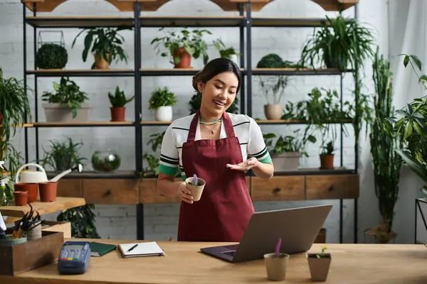 A young Asian woman, the owner of a small plant shop, smiles while showing off new plants to her customers. — Stock Photo