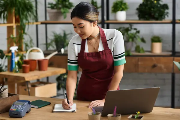 Une jeune femme asiatique dans un tablier rouge travaille dans son magasin de plantes, faisant l'inventaire ou gérant des commandes. — Photo de stock