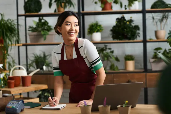 An Asian woman wearing an apron smiles while taking inventory in her plant shop. — Stock Photo