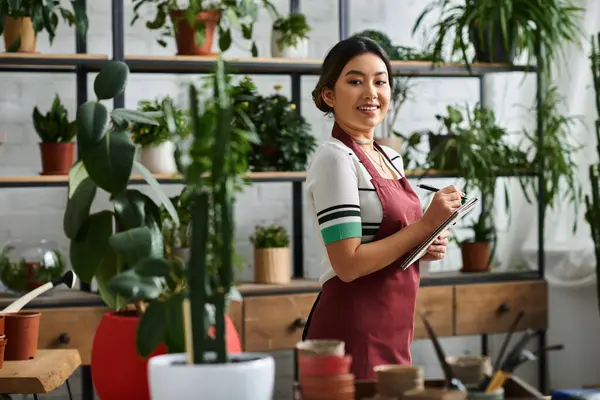 Eine junge Frau in Schürze steht in ihrem Pflanzenladen, macht Inventur und ist von üppigem Grün umgeben.. — Stockfoto