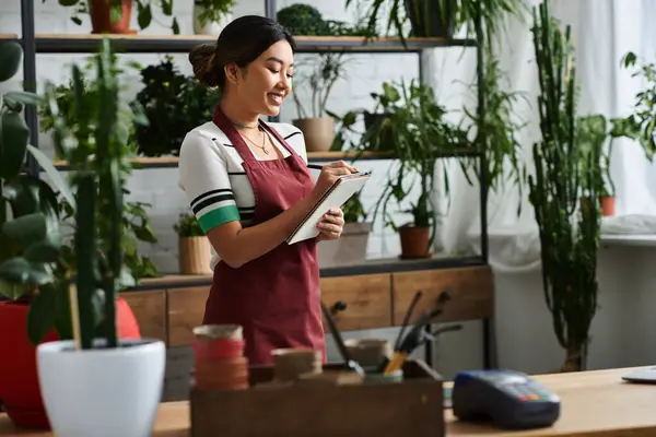 A young Asian woman, wearing an apron, smiles as she takes inventory in her plant shop. — Stock Photo