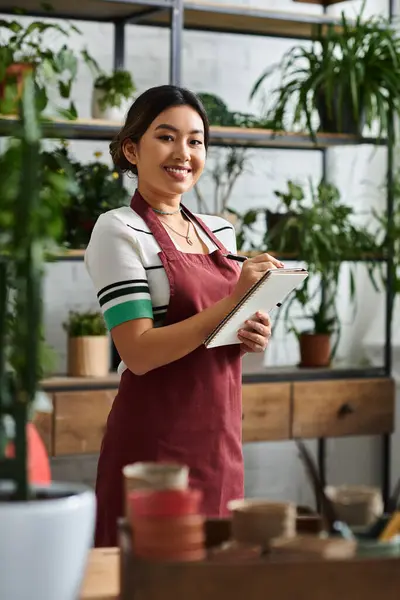 An Asian woman in an apron takes inventory in her plant shop, surrounded by vibrant greenery. — Stock Photo