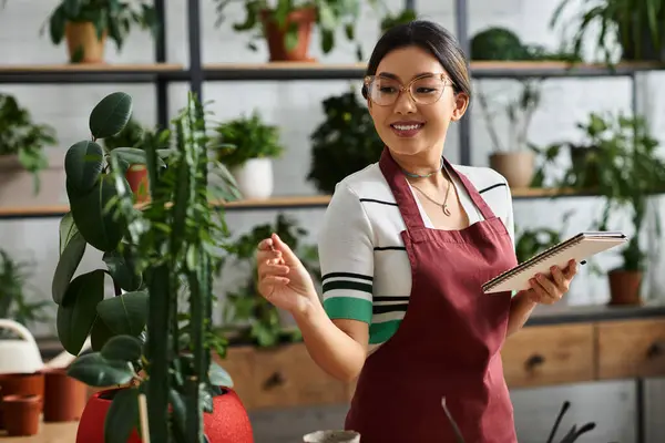 An Asian woman wearing a red apron stands in her plant shop, surrounded by greenery. She is checking her notes while smiling. — Stock Photo