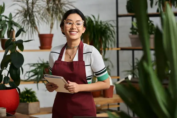 Eine glückliche Asiatin, Inhaberin eines Pflanzengeschäfts, steht in ihrem Geschäft, ein Notizbuch in der Hand, umgeben von üppigem Grün. — Stockfoto