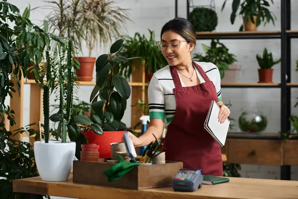 Una mujer asiática sonriente, con un delantal, trabaja en su tienda de plantas, rodeada de exuberante vegetación. - foto de stock