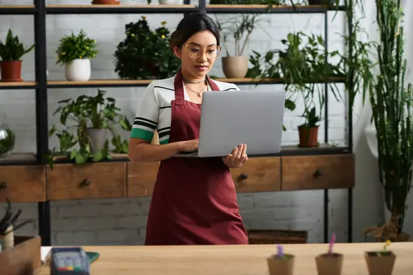 A young Asian woman, wearing an apron, stands in her plant store and uses her laptop to manage her business online. — Stock Photo