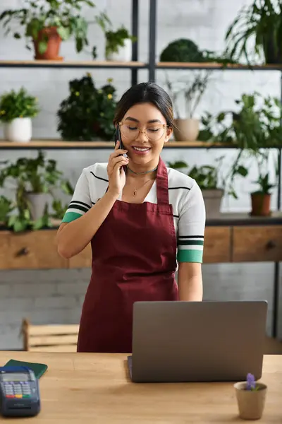 Una mujer asiática en un delantal sonríe mientras recibe una llamada en su tienda de plantas con una computadora portátil y una caja registradora cerca. - foto de stock