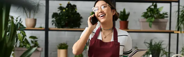 Une jeune femme dans un tablier sourit alors qu'elle prend un appel téléphonique alors qu'elle travaille dans son atelier d'usine. — Photo de stock