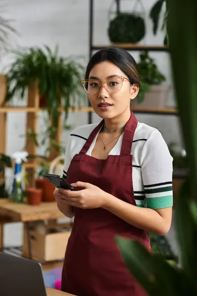 An Asian woman, wearing an apron, stands in her plant shop and looks at her phone, surrounded by lush greenery. — Stock Photo