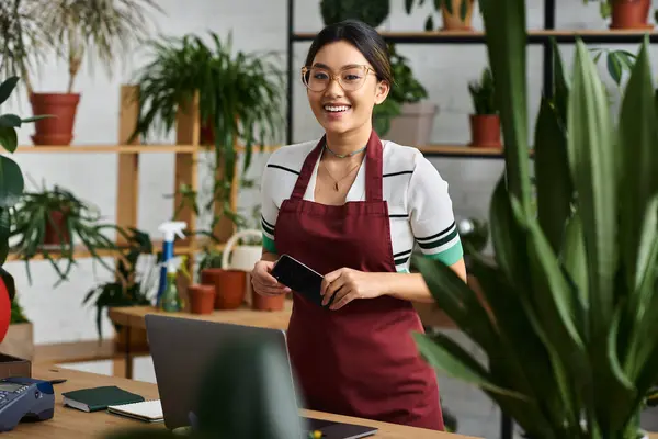 A young Asian woman in a red apron smiles while working in her plant shop, surrounded by greenery. — Stock Photo