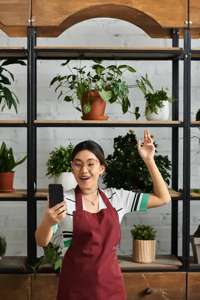 An Asian woman, the owner of a plant shop, enthusiastically greets her customers with a smile and a wave. — Stock Photo