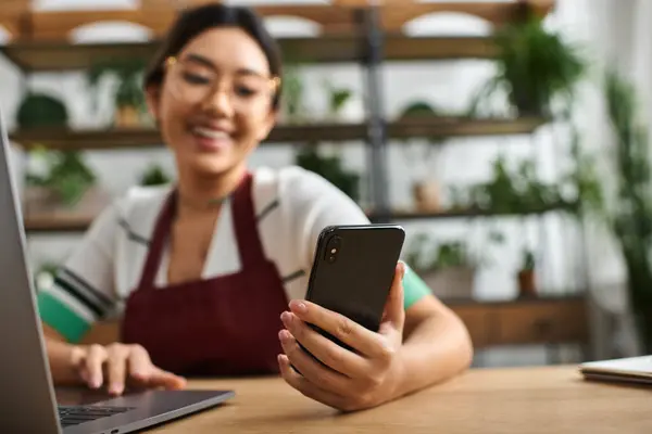 A young Asian woman in an apron uses her phone while working in her plant store. — Stock Photo