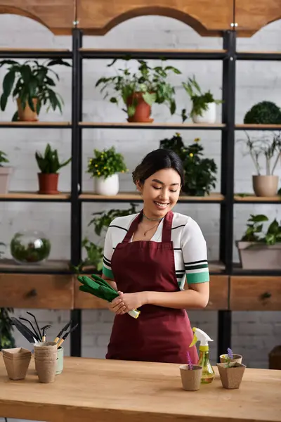 A young woman wearing a red apron smiles as she works in her plant shop, surrounded by lush greenery. — Stock Photo