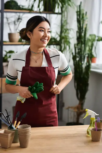 Une femme asiatique souriante, portant un tablier et tenant des gants de jardinage, se tient dans son magasin de plantes, prête à aider les clients. — Photo de stock