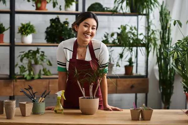 A young Asian woman, the owner of a plant shop, smiles brightly as she works amongst her thriving collection of greenery. — Stock Photo