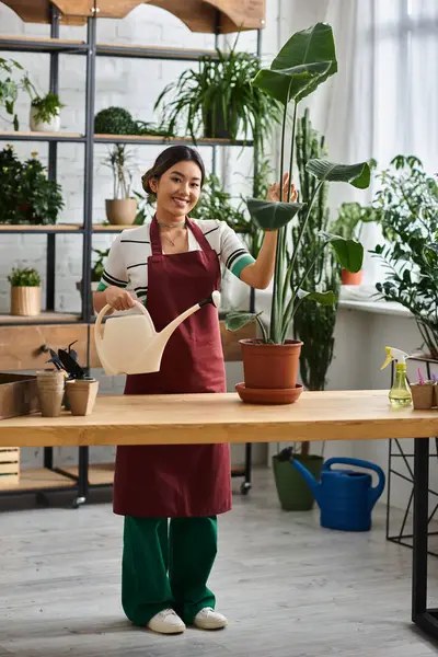 A young Asian woman, the proud owner of a plant shop, waters a large houseplant with a gentle smile. — Stock Photo