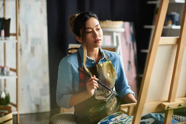 An Asian artist in an apron stands in her studio, thoughtfully considering her next brushstroke on a canvas. — Stock Photo