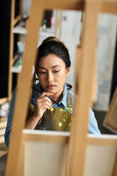 A young Asian artist, wearing an apron, thoughtfully paints in her workshop. — Stock Photo