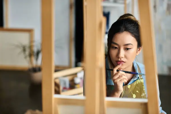 A young Asian artist, wearing an apron, thoughtfully considers her next brushstroke in her workshop. — Stock Photo