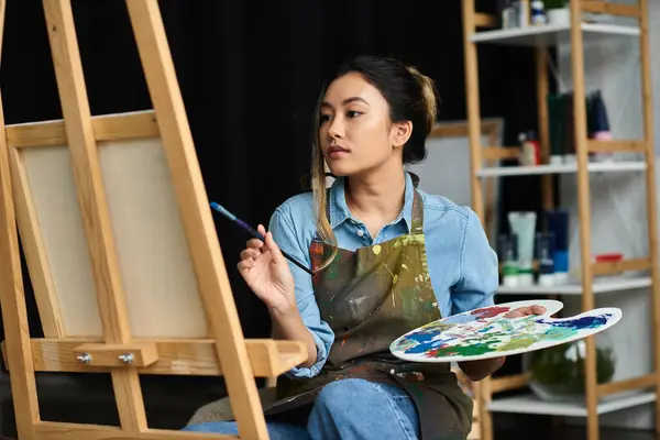 An Asian artist in an apron sits in her workshop, holding a paintbrush and a palette, focused on her art. — Stock Photo