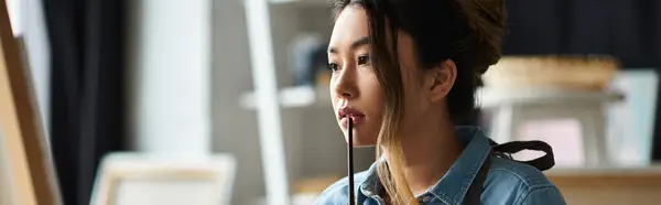 A young Asian artist in an apron ponders her next brushstroke in her workshop. — Stock Photo