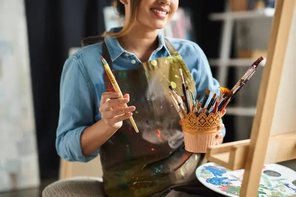A young Asian woman artist, wearing an apron, holds a paintbrush and smiles while working in her art studio. — Stock Photo