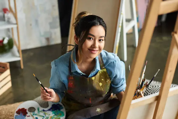 A young Asian woman artist smiles while holding a paintbrush and standing in front of an easel in her art studio. — Stock Photo