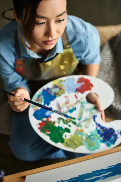 A young Asian woman in an apron holds a palette of paints, concentrating on her art in a workshop. — Stock Photo