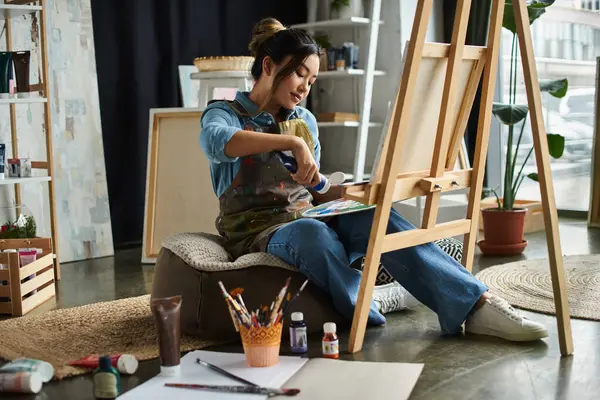 Eine junge asiatische Künstlerin mit Schürze sitzt auf einem Kissen in ihrer Werkstatt und konzentriert sich auf ihre Leinwand und Farbe. — Stockfoto